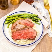 Sliced Air Fryer Eye of Round Roast on mashed potatoes with asparagus on a plate, set on a table with a napkin and utensils.
