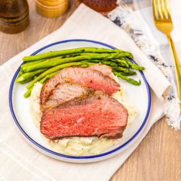 Sliced Air Fryer Eye of Round Roast on mashed potatoes with asparagus on a plate, set on a table with a napkin and utensils.