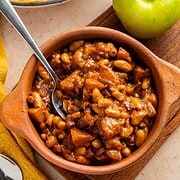 Apple Pie Baked Beans in a brown bowl with a spoon, next to a green apple on a wooden board.