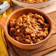 A bowl of Apple Pie Baked Beans in a rich brown sauce, placed on a wooden board with a yellow cloth nearby.