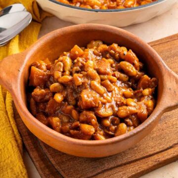A bowl of Apple Pie Baked Beans in a rich brown sauce, placed on a wooden board with a yellow cloth nearby.