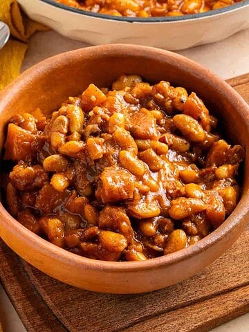A bowl of Apple Pie Baked Beans in a rich brown sauce, placed on a wooden board with a yellow cloth nearby.