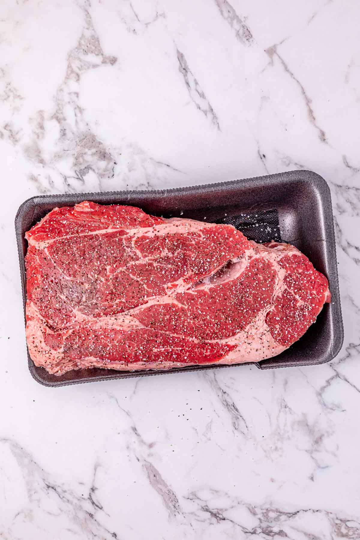 Raw seasoned beef steak in a black plastic tray on a white marble surface, perfect for preparing a flavorful Slow Cooker French Onion Pot Roast.