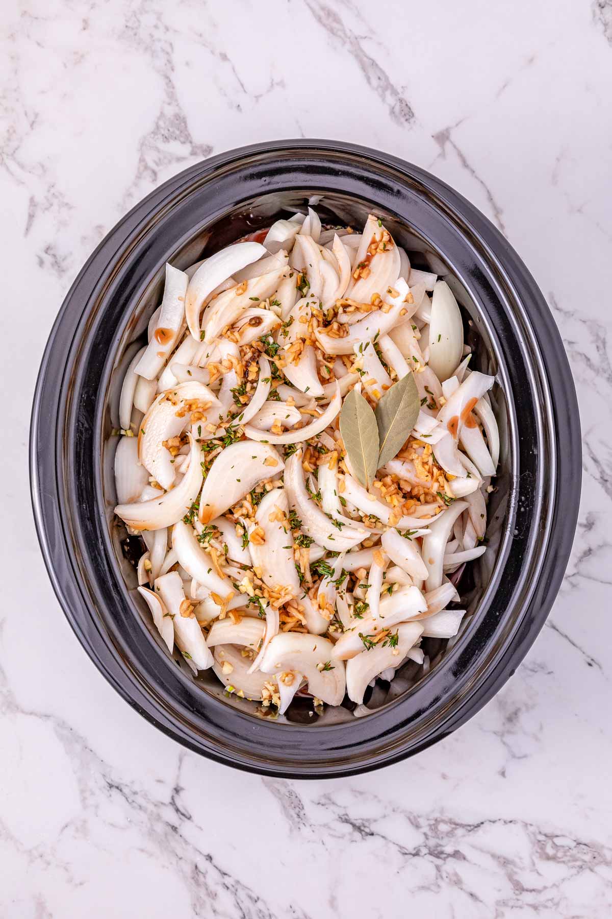 Sliced onions, garlic, spices, and bay leaves in a slow cooker on a marble countertop—perfect beginnings for a flavorful Slow Cooker French Onion Pot Roast.