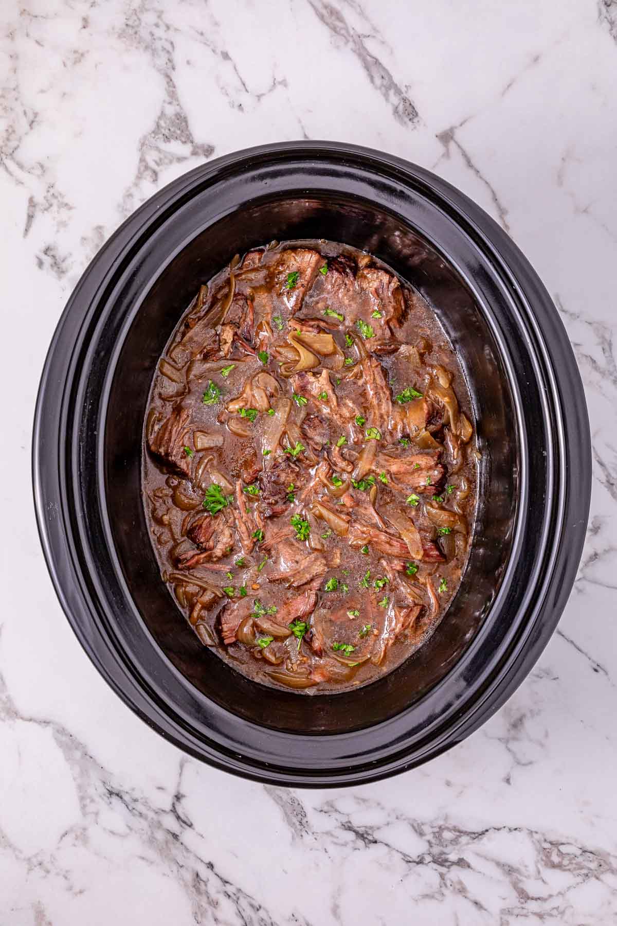 Slow Cooker French Onion Pot Roast: Shredded beef with onions and sauce in a black slow cooker, garnished with parsley on a marble countertop.