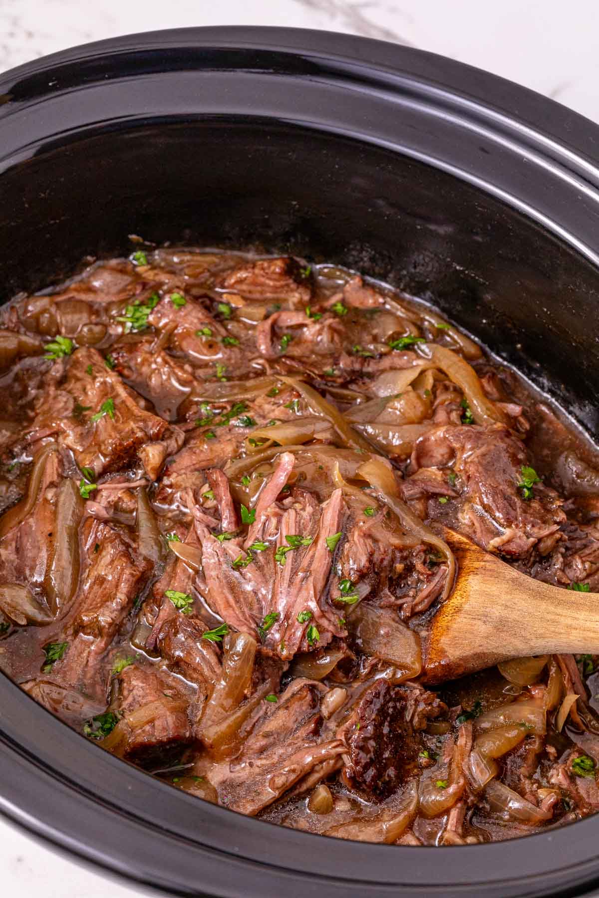 Shredded beef with onions and sauce in a black slow cooker, stirred with a wooden spoon—a delicious Slow Cooker French Onion Pot Roast.