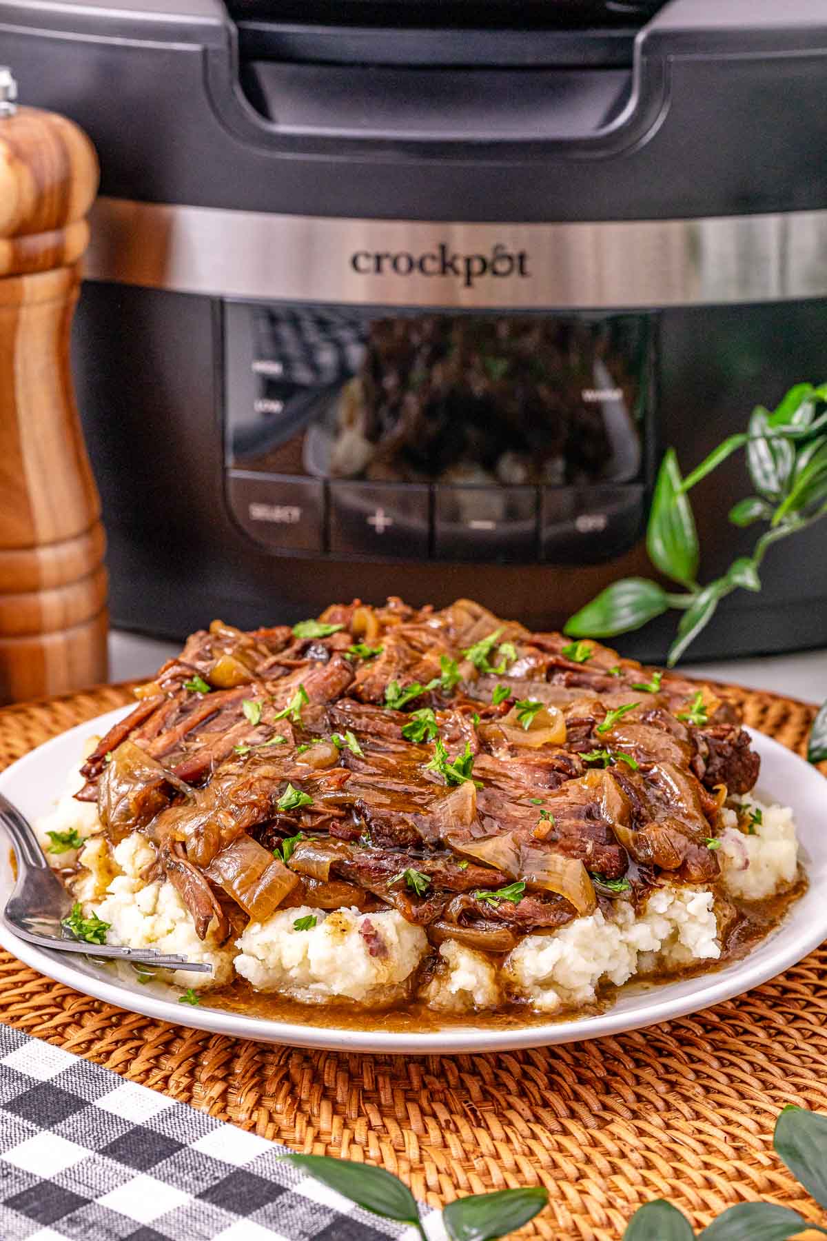A plate of Slow Cooker French Onion Pot Roast with onions and rich gravy over mashed potatoes, with a crockpot in the background.