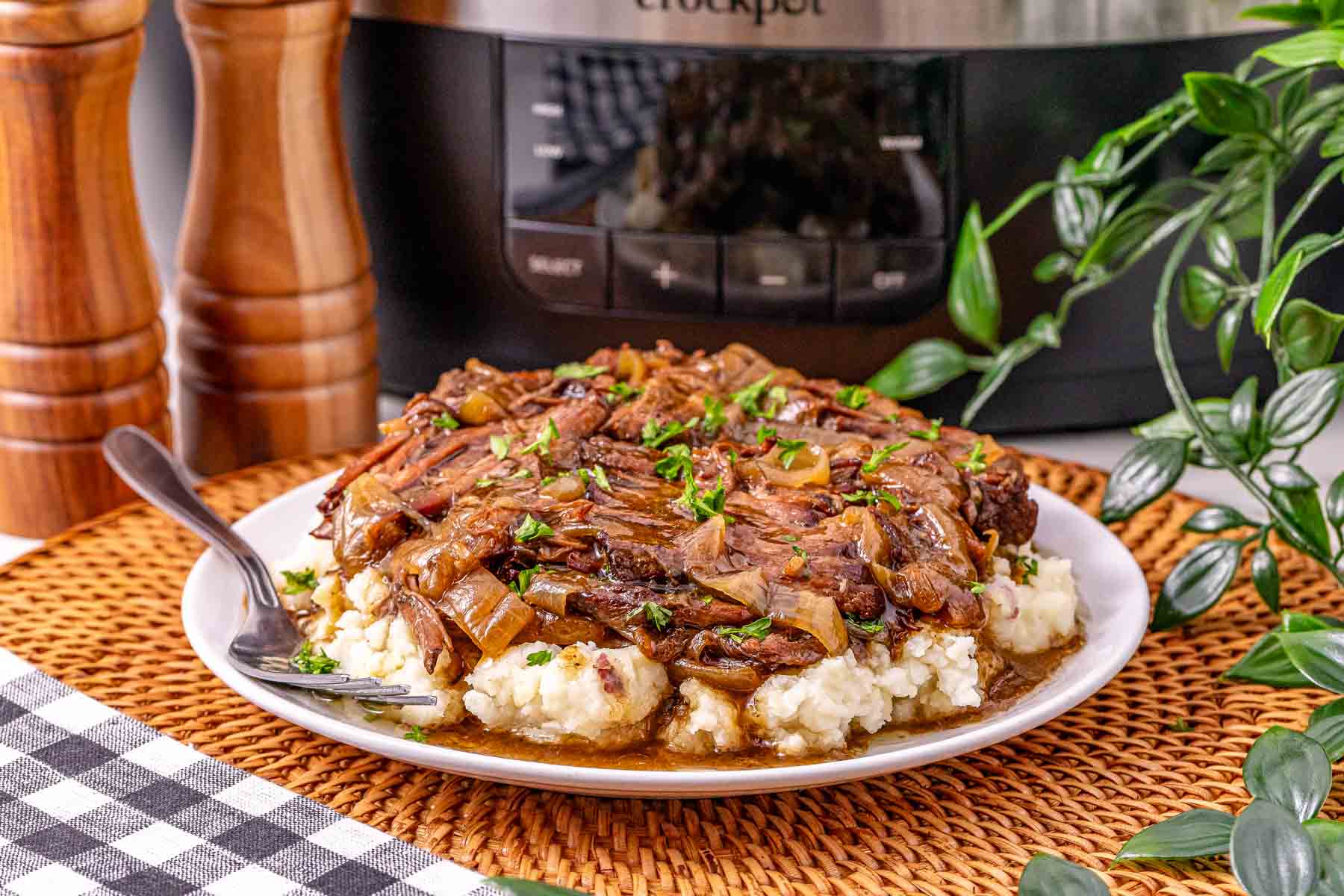 Slow Cooker French Onion Pot Roast with rich onions and savory gravy served over creamy mashed potatoes on a plate, nestled beside a slow cooker and surrounded by fresh plants.