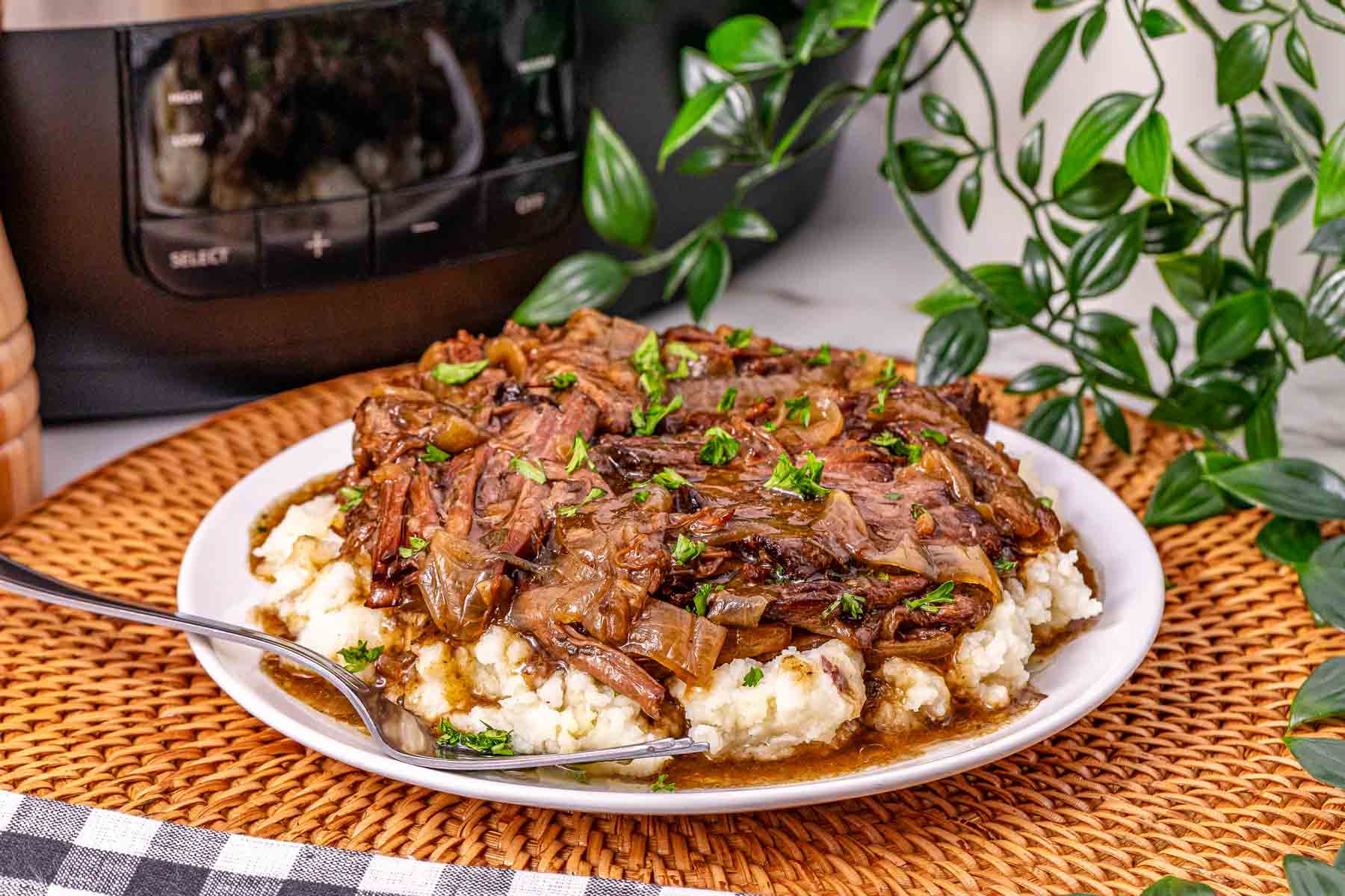 Slow Cooker French Onion Pot Roast shredded beef with onions atop creamy mashed potatoes, served on a white plate with a fork, plants in the background.