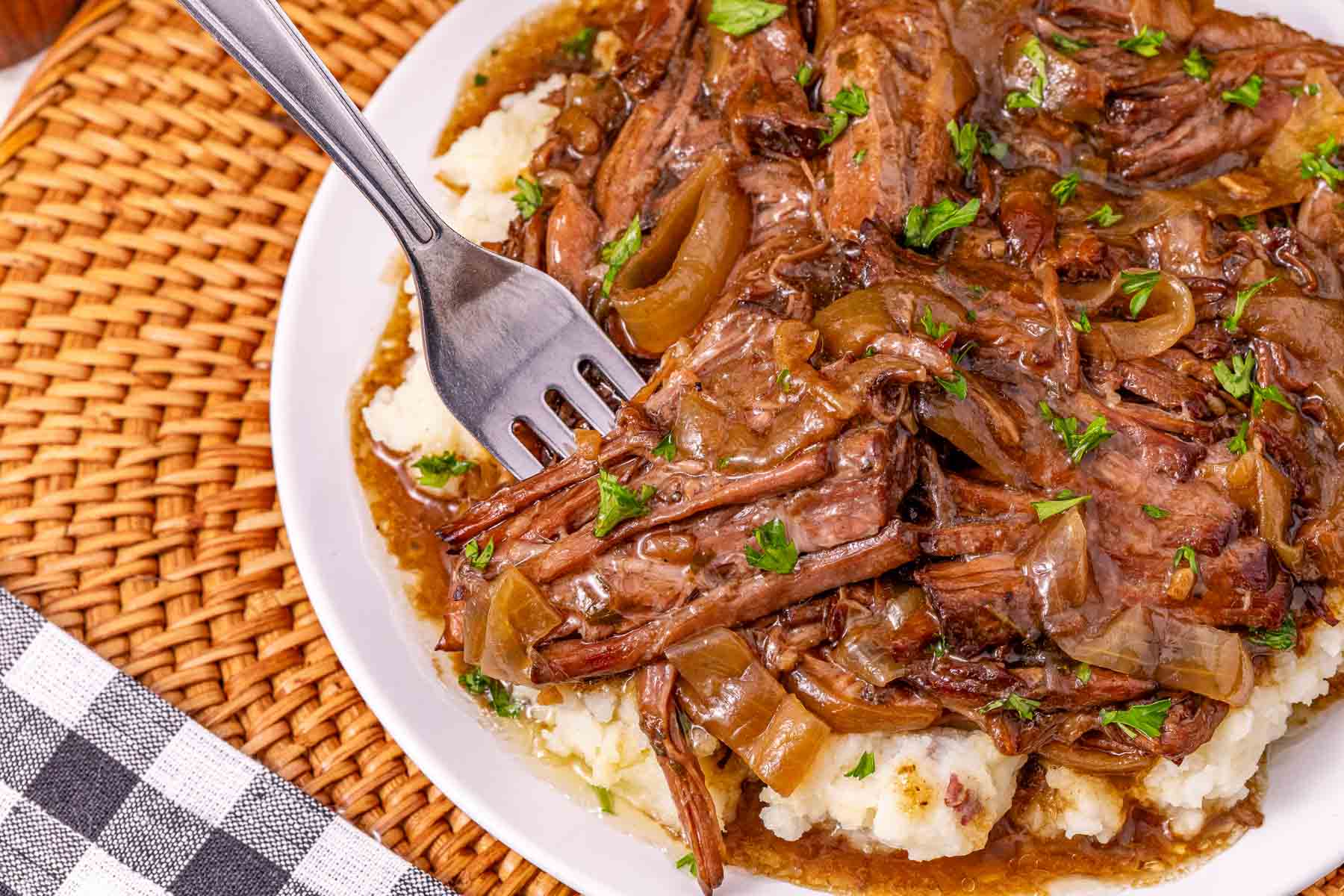 Slow Cooker French Onion Pot Roast—fork shredding pot roast with savory onions and rich gravy over mashed potatoes on a white plate.