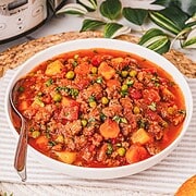 Bowl of hearty Crockpot Poor Man’s Stew with beef, carrots, peas, and potatoes in a rich tomato broth, placed beside a slow cooker.
