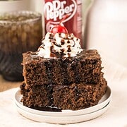 Two Dr. Pepper Brownies topped with whipped cream, a cherry, and chocolate drizzle, with a Dr Pepper can in the background.