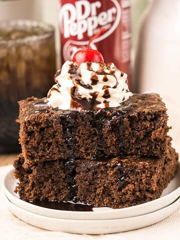 Two Dr. Pepper Brownies topped with whipped cream, a cherry, and chocolate drizzle, with a Dr Pepper can in the background.