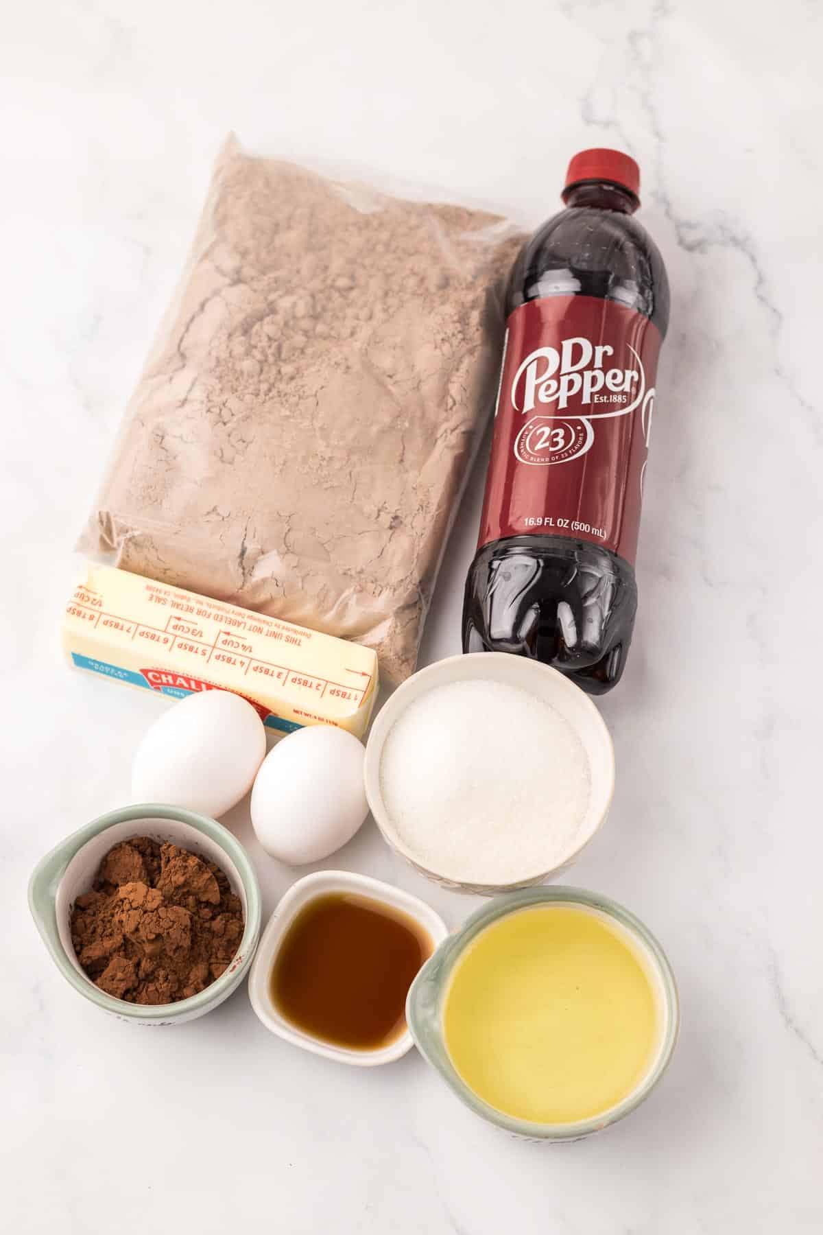Ingredients for Dr. Pepper Brownies are laid out on a white surface: cake mix, a Dr Pepper bottle, two eggs, butter, cocoa, vanilla, oil, and sugar.