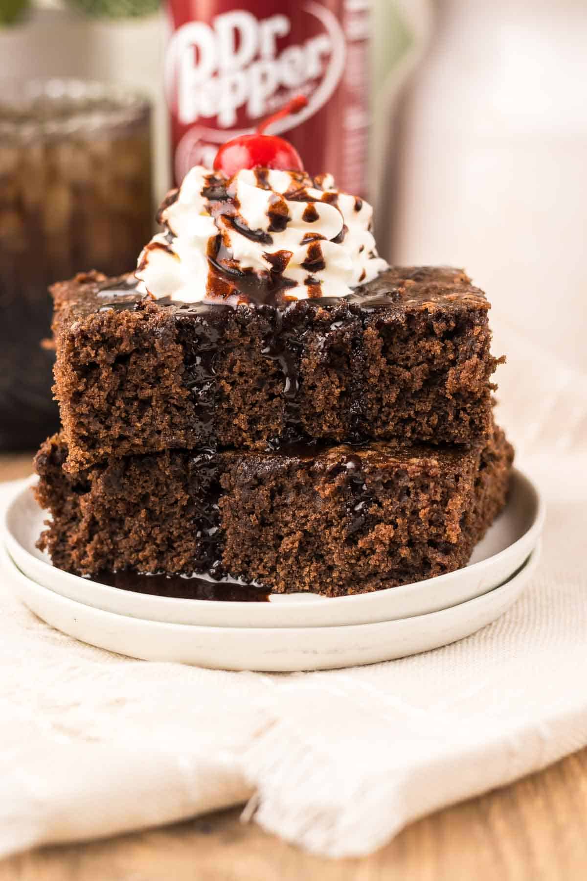 Two stacked chocolate cake slices topped with whipped cream, a rich chocolate drizzle, and a cherry on a white plate evoke the decadent appeal of Dr. Pepper Brownies.