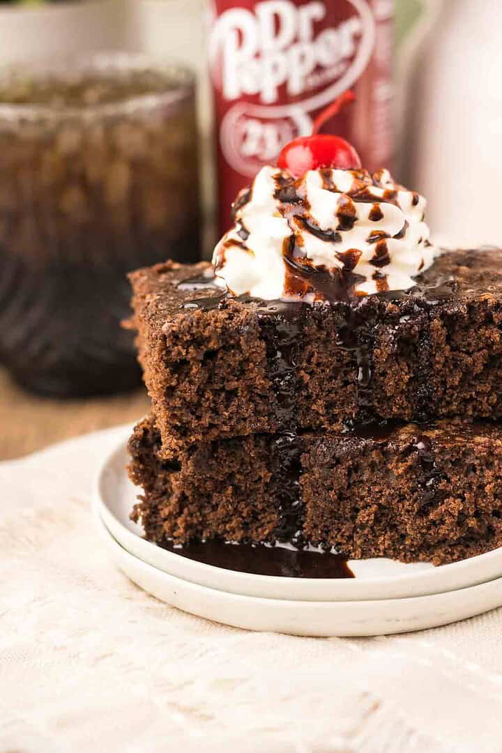 Two pieces of chocolate cake, reminiscent of rich Dr. Pepper Brownies, are served with fluffy whipped cream, chocolate syrup, and a cherry on top, with a drink in the background.