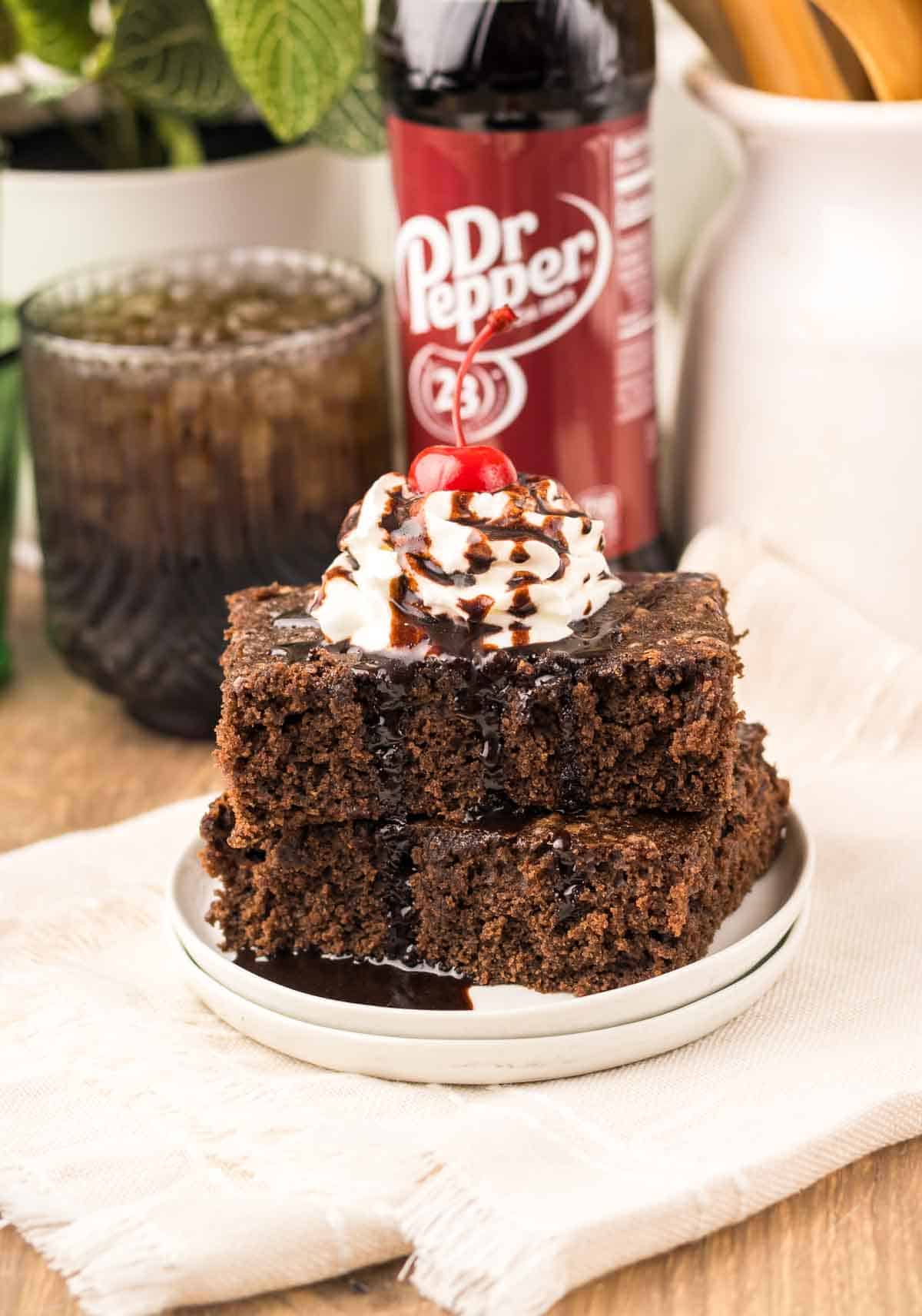 Two decadent chocolate cake slices topped with whipped cream, chocolate syrup, and a cherry are served with a Dr Pepper bottle in the background—perfectly reminiscent of classic Dr. Pepper Brownies for a rich and flavorful treat.
