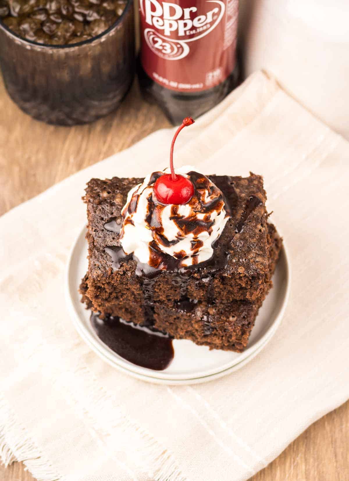A slice of chocolate cake with whipped cream, chocolate syrup, and a cherry on top, reminiscent of rich Dr. Pepper Brownies, is served on a small white plate.
