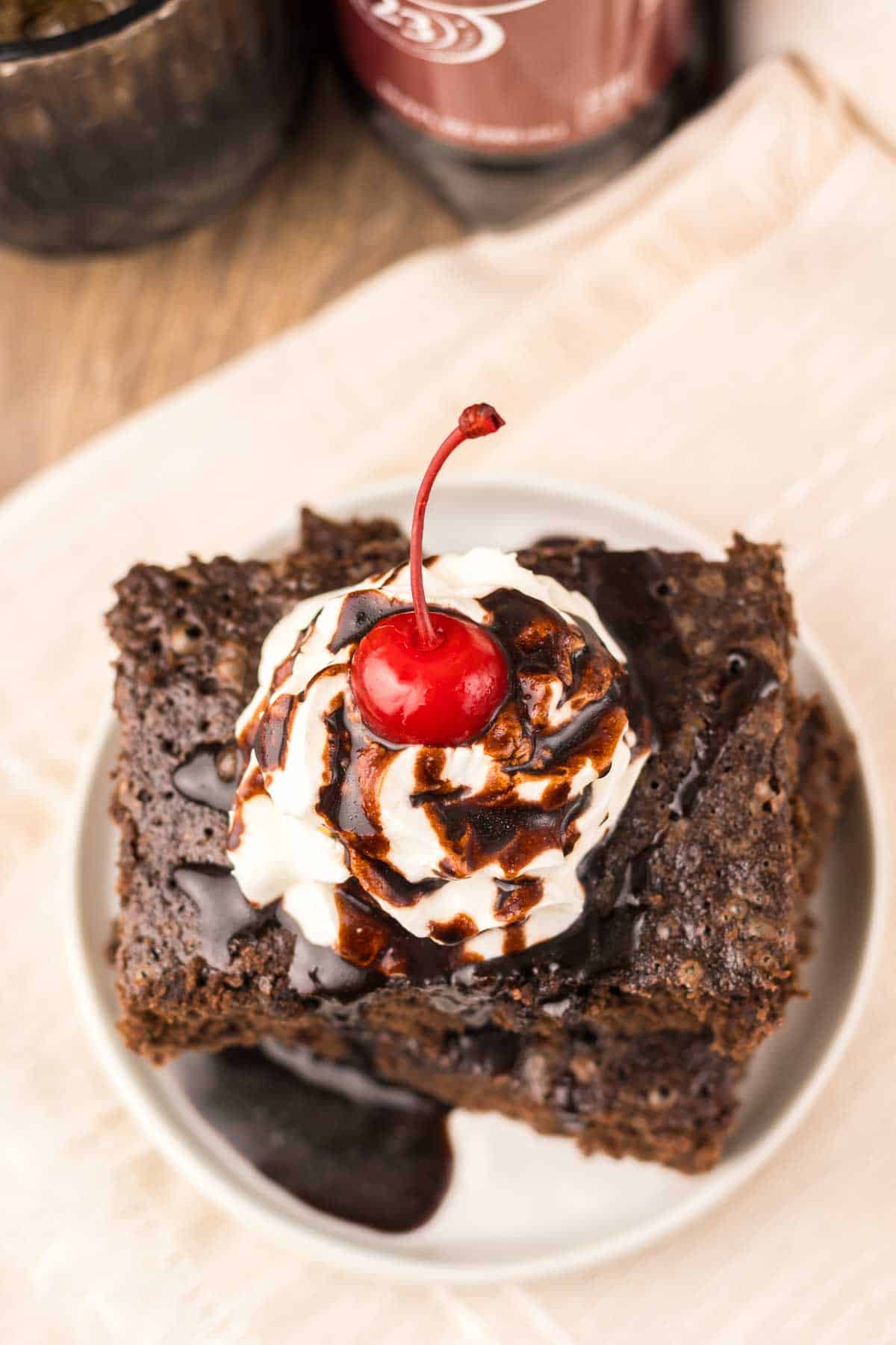 Dr. Pepper Brownies topped with whipped cream, chocolate syrup, and a cherry on a white plate.