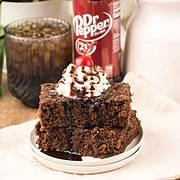 Two chocolate cake squares with whipped cream and cherry, reminiscent of Dr. Pepper Brownies, sit next to a Dr Pepper bottle and a glass of soda.