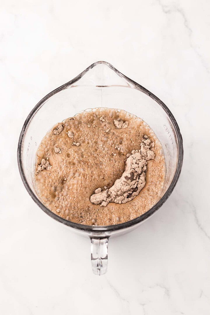 A glass measuring cup with bubbly brown yeast mixture, reminiscent of the rich hue found in Dr. Pepper Brownies, and dry yeast on a white marble surface.