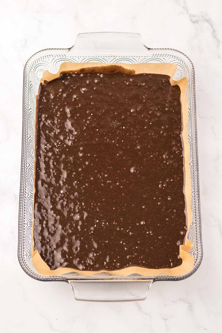 A glass baking dish with parchment paper filled with rich Dr. Pepper Brownies batter, ready to be baked.
