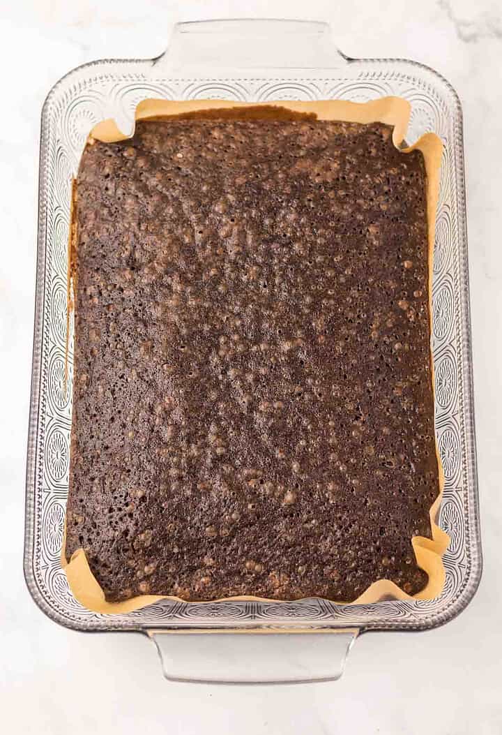 A rectangular baking dish lined with parchment paper contains a large, uncut Dr. Pepper Brownie, its rich chocolate surface hinting at the unique flavor twist inside.