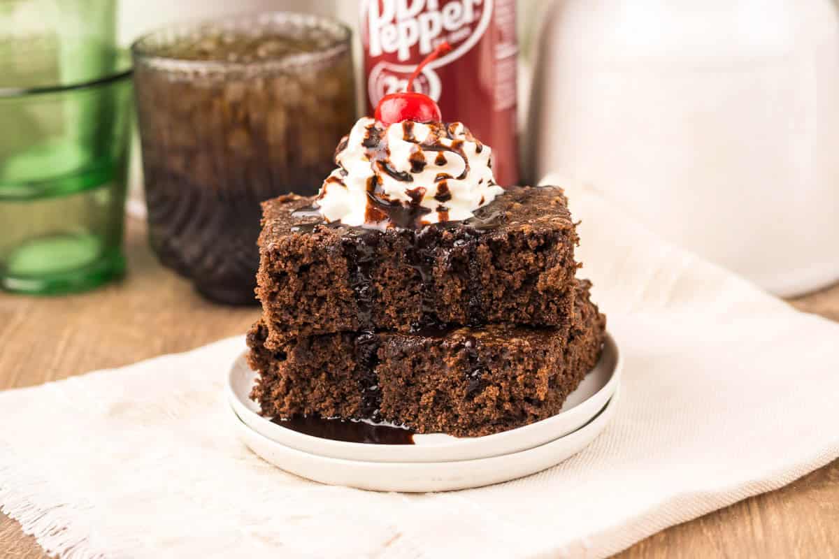 Two Dr. Pepper Brownies stacked, topped with whipped cream, chocolate sauce, and a cherry, on a white plate.