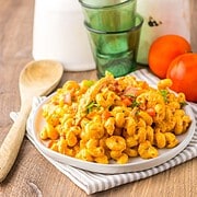 Plate of creamy pasta with vegetables on a striped napkin, wooden spoons, and tomatoes in the background—this dish brings to mind the hearty flavors of slow cooker cheesy chicken enchilada pasta, blending comfort food vibes with fresh ingredients.