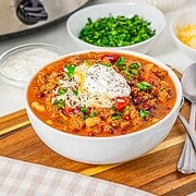 Bowl of Slow Cooker Chili with Sausage topped with cheese, sour cream, and herbs, with sides and a slow cooker in the background.