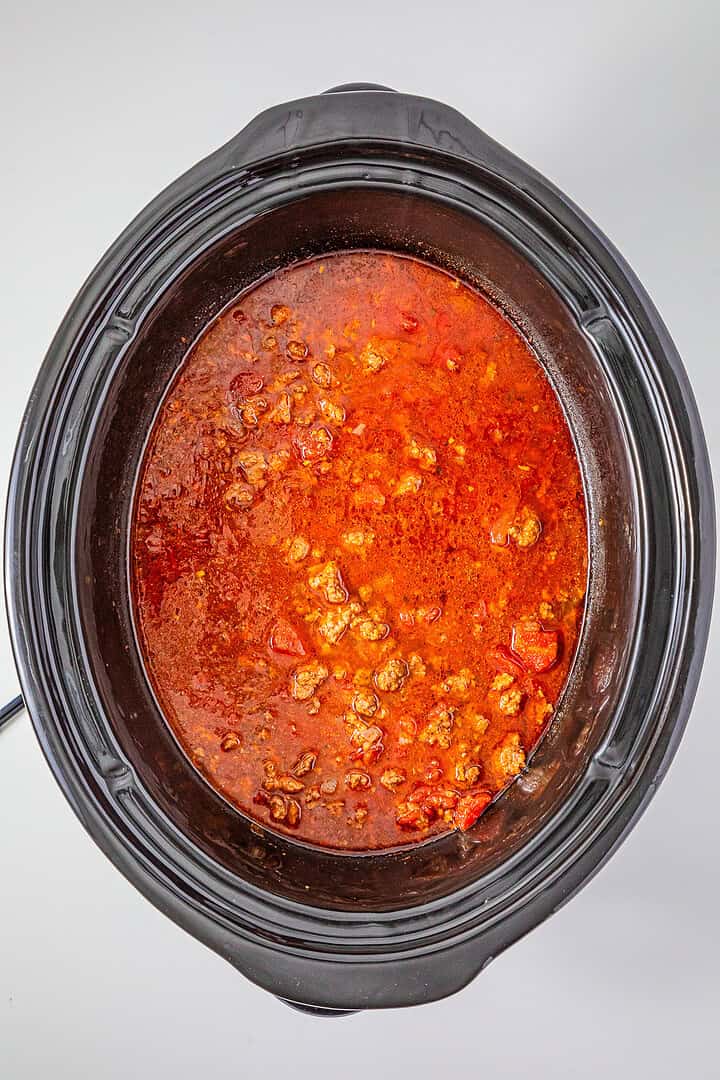 A slow cooker filled with red, chunky Slow Cooker Chili with Sausage sits on a white surface.
