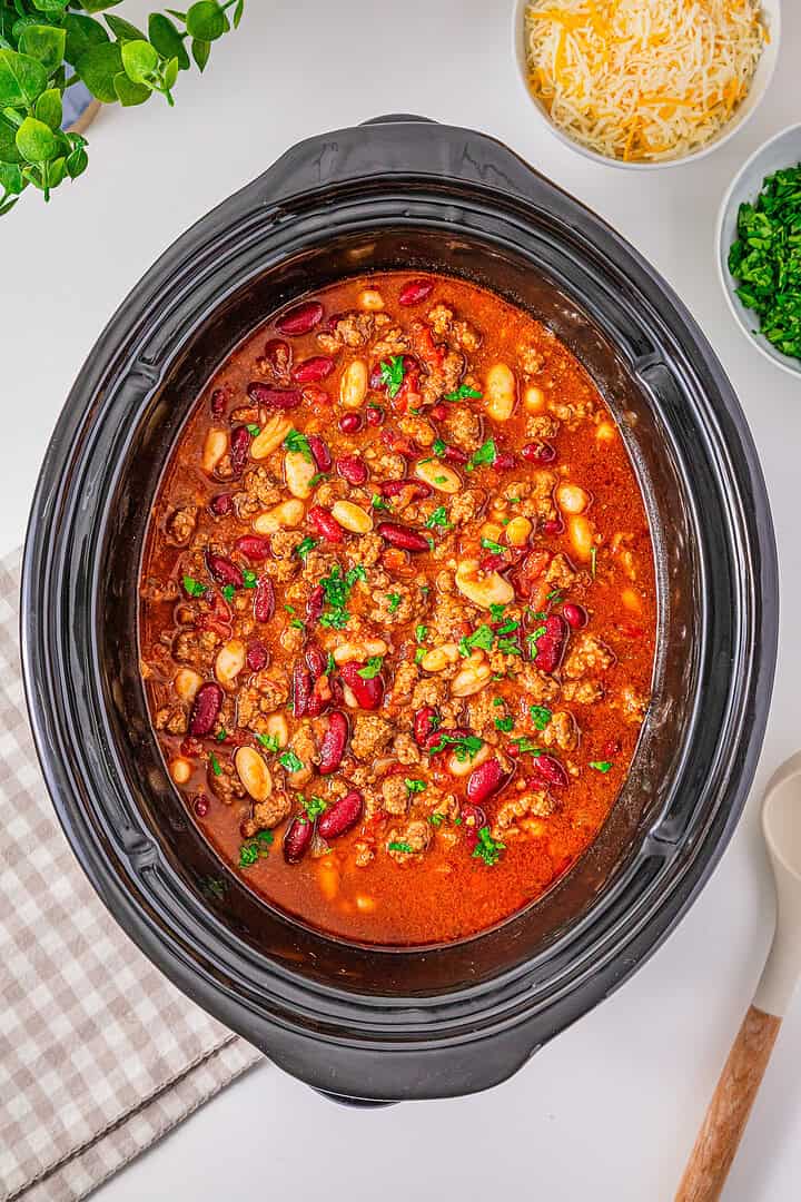 Slow Cooker Chili with Sausage simmers in the slow cooker, topped with fresh herbs, with cheese and chopped parsley in bowls nearby.