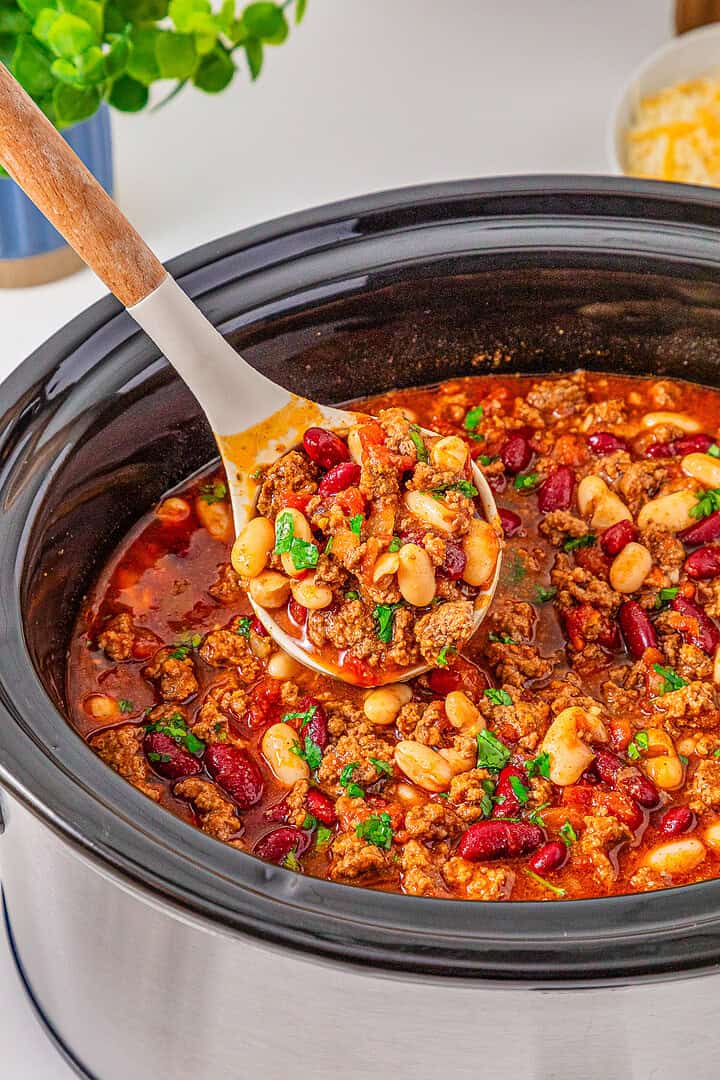 A ladle scoops hearty Slow Cooker Chili with Sausage, beans, and ground meat from a slow cooker, garnished with herbs.
