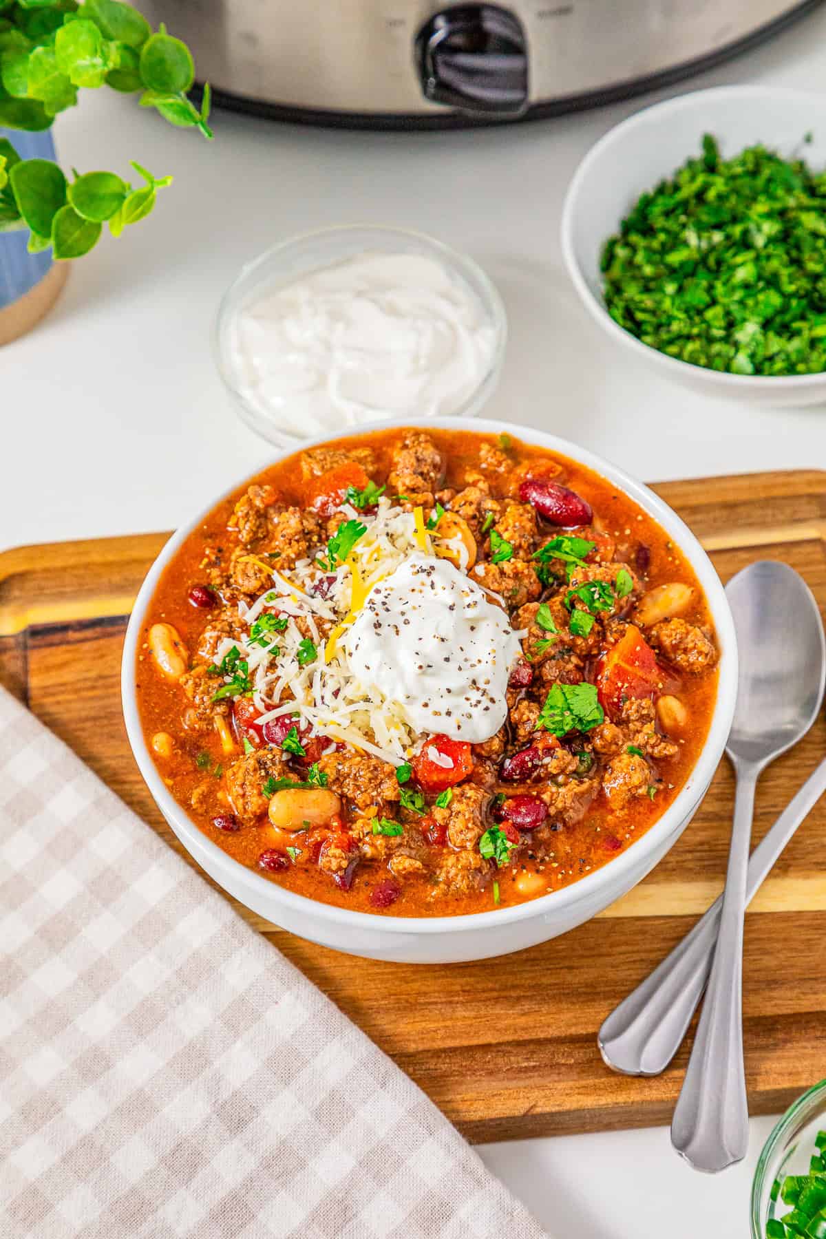 A bowl of Slow Cooker Chili with Sausage topped with sour cream, cheese, and herbs sits on a wooden board alongside spoons and a napkin.
