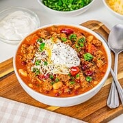 Bowl of Slow Cooker Chili with Sausage, beans, ground meat, cheese, sour cream, and herbs, next to spoon and napkin.