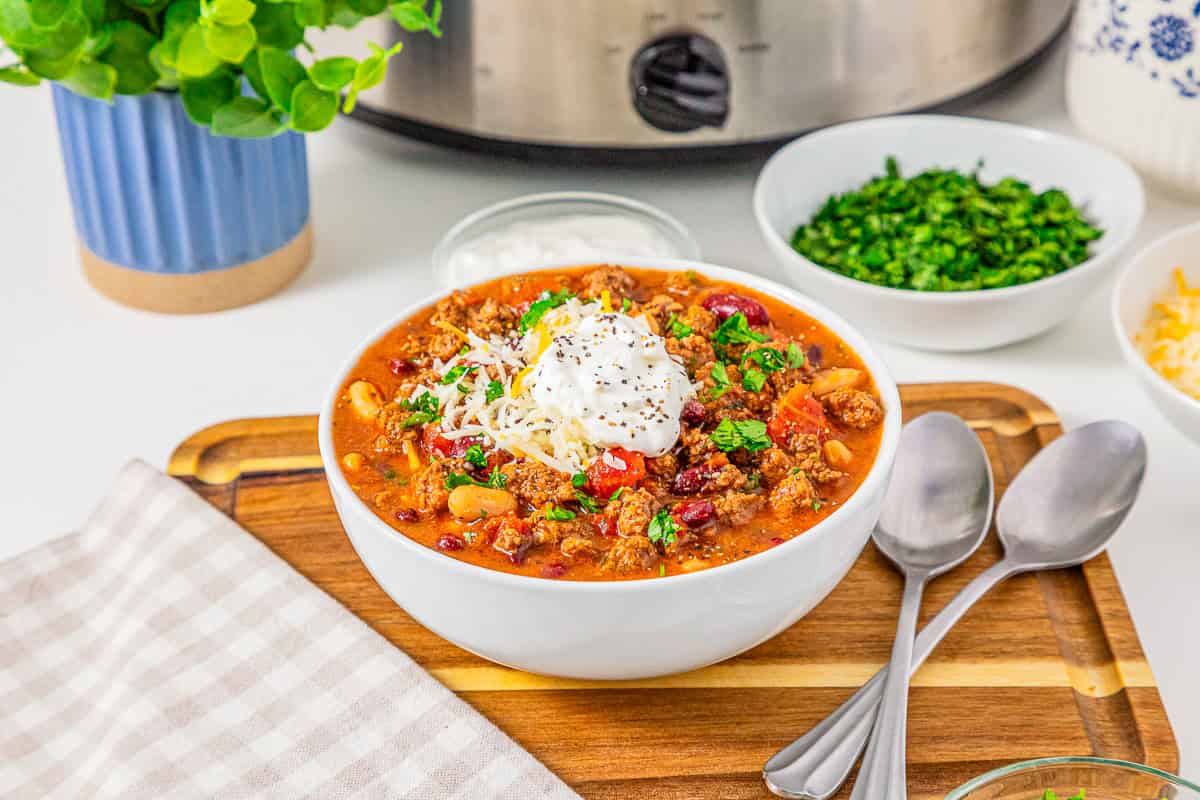 A hearty bowl of Slow Cooker Chili with Sausage topped with sour cream, cheese, and herbs, served alongside spoons and extra toppings on a rustic wooden tray.