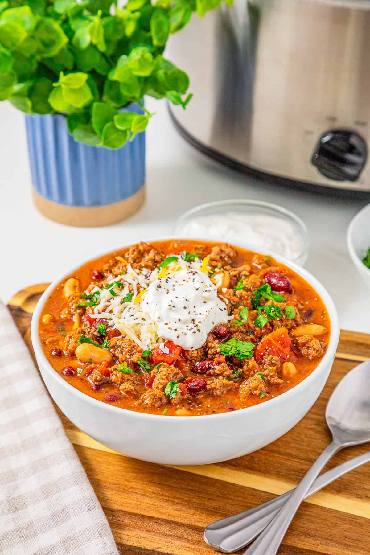 Bowl of slow cooker chili with sausage topped with sour cream, cheese, and herbs, served with a spoon and napkin on a wooden board.