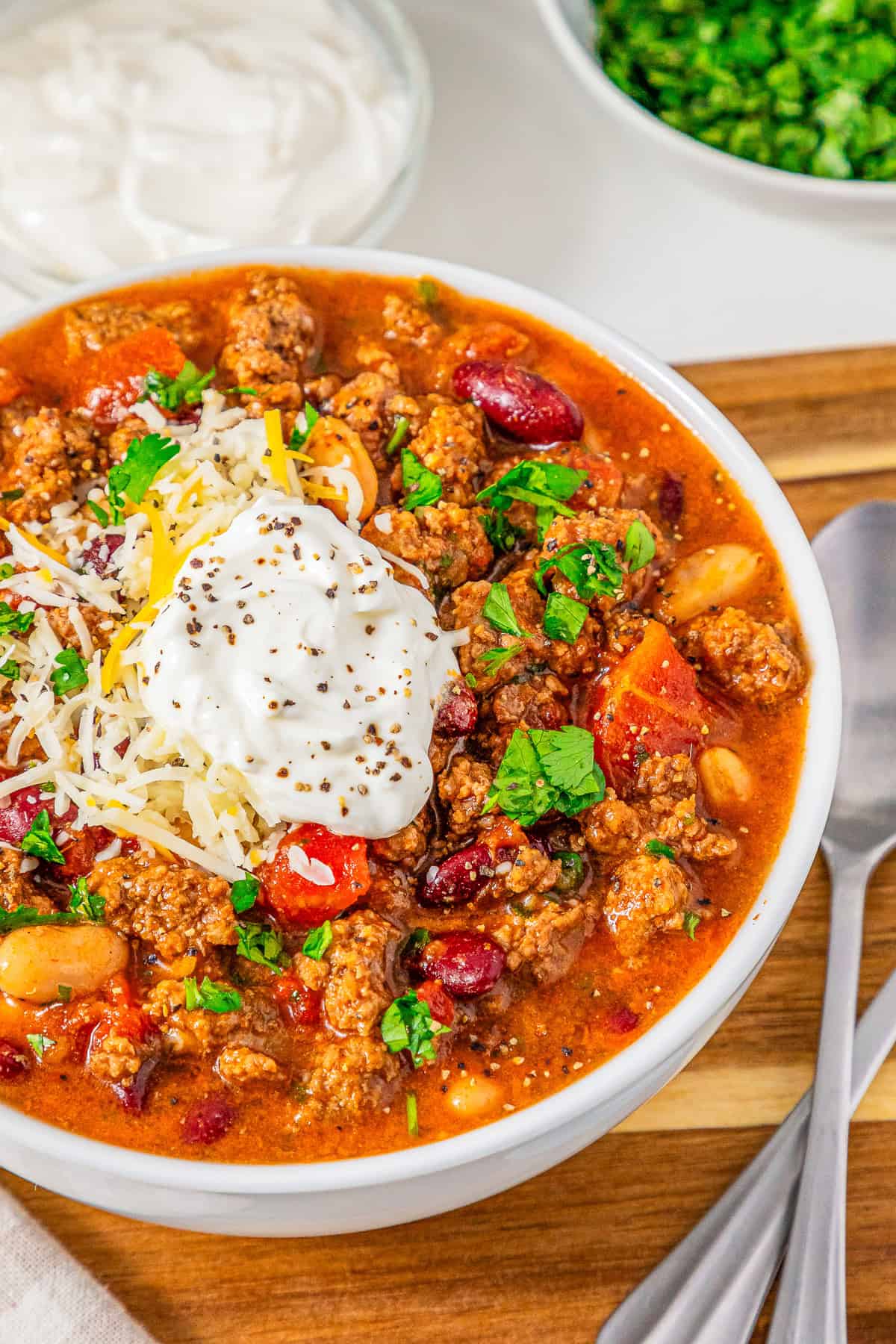 Bowl of Slow Cooker Chili with Sausage topped with shredded cheese, sour cream, and chopped herbs, with spoons beside it.