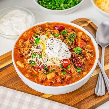 Bowl of Slow Cooker Chili with Sausage, beans, ground meat, cheese, sour cream, and herbs, next to spoon and napkin.