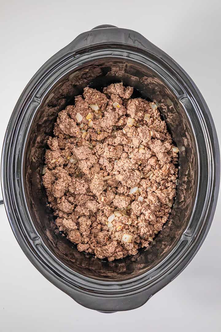 Ground beef and chopped onions cooking in a slow cooker, viewed from above on a white background—perfect ingredients for starting a flavorful Slow Cooker Chili with Sausage.
