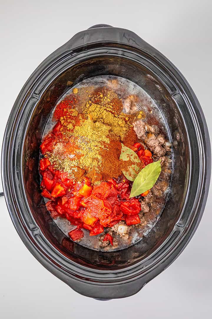 Overhead view of a slow cooker with ground beef, sausage, diced tomatoes, spices, and bay leaves, ready to be mixed for Slow Cooker Chili with Sausage.