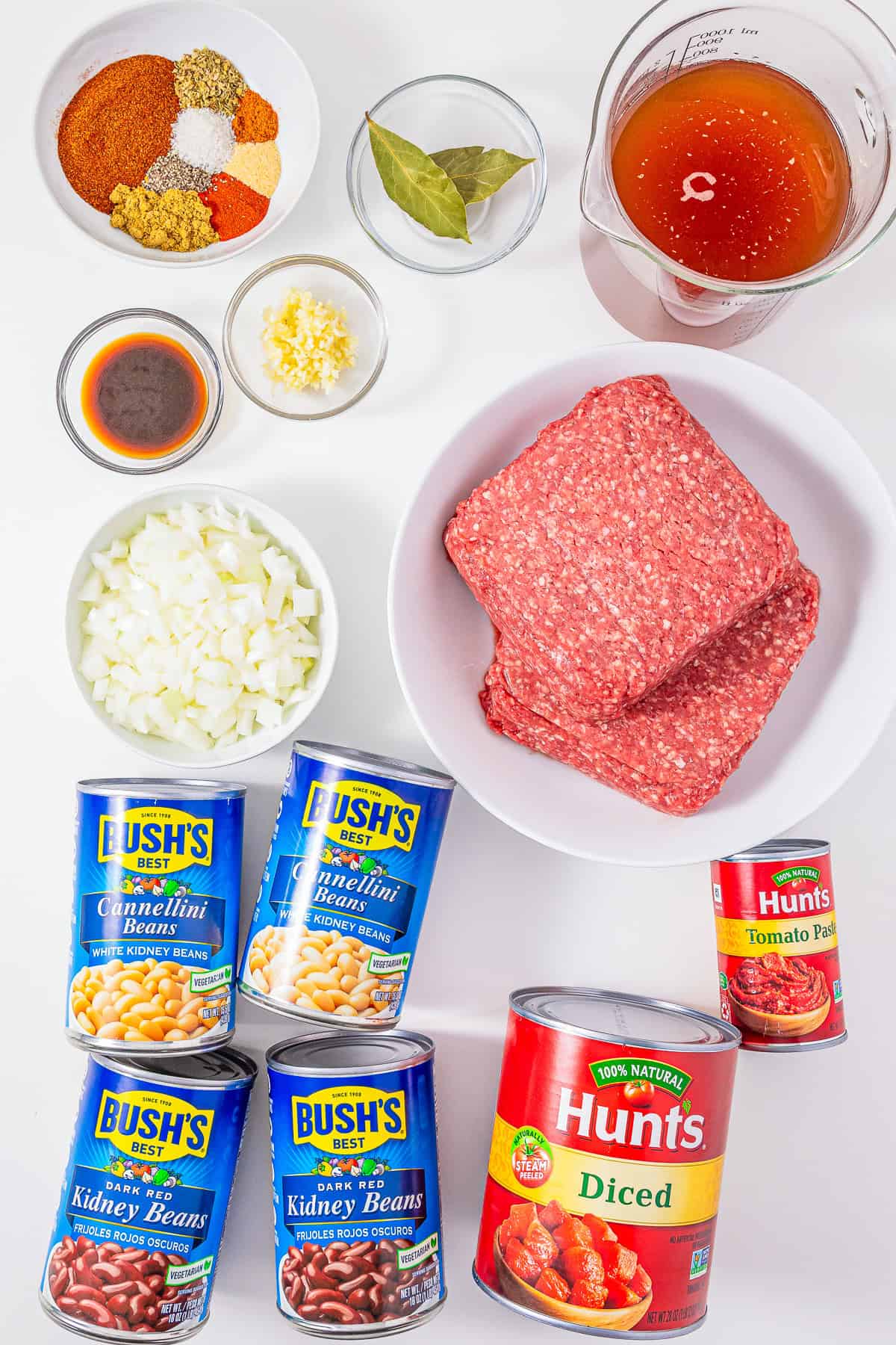 Chopped onions, garlic, ground beef, sausage, spices, broth, beans, tomato paste, and diced tomatoes on a white surface—perfect ingredients for making hearty Slow Cooker Chili with Sausage.