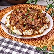Shredded beef with gravy over mashed potatoes on a white plate, featuring the rich flavors of Slow Cooker French Onion Pot Roast, with a crockpot and greenery in the background.