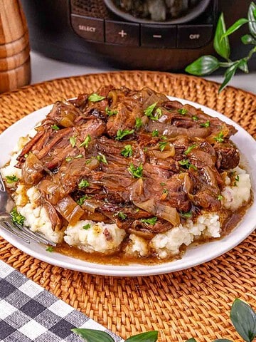 Slow Cooker French Onion Pot Roast features tender braised beef with caramelized onions and rich gravy, served over creamy mashed potatoes and garnished with parsley on a white plate.