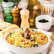 A bowl of couscous with vegetables and sausage, reminiscent of flavors found in Slow Cooker Italian Wedding Soup, set on a table with bread, glassware, and condiments.