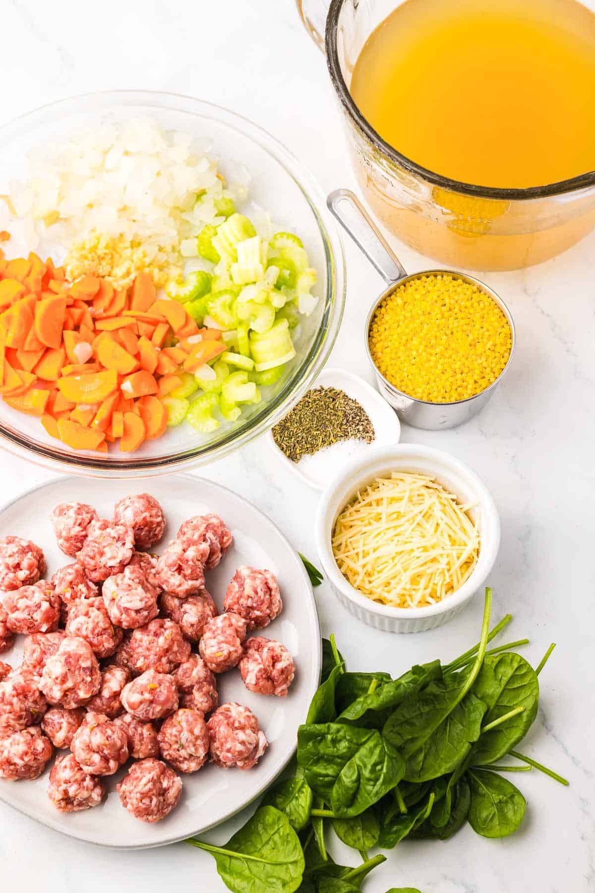 Bowls of chopped onion, carrot, celery, raw sausage, spinach, cheese, broth, dry pasta, and herbs on a white surface—everything you need to create a hearty Slow Cooker Italian Wedding Soup.