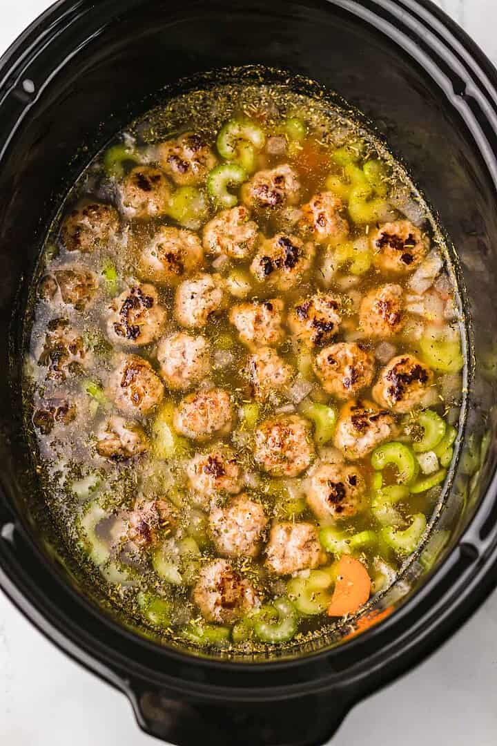Meatballs, celery, and broth simmering in a black slow cooker create the delicious base for Slow Cooker Italian Wedding Soup, viewed from above.