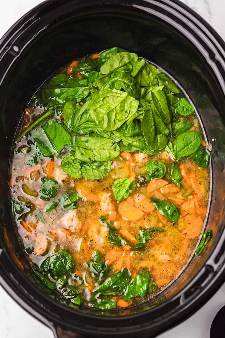 Spinach, carrots, and broth simmer together in a black slow cooker for a comforting Slow Cooker Italian Wedding Soup, with fresh spinach leaves layered on top.