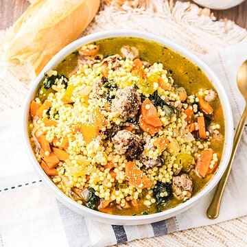 A bowl of Slow Cooker Italian Wedding Soup featuring tender meatballs, carrots, fresh greens, and couscous, served with crusty bread and a gold spoon.
