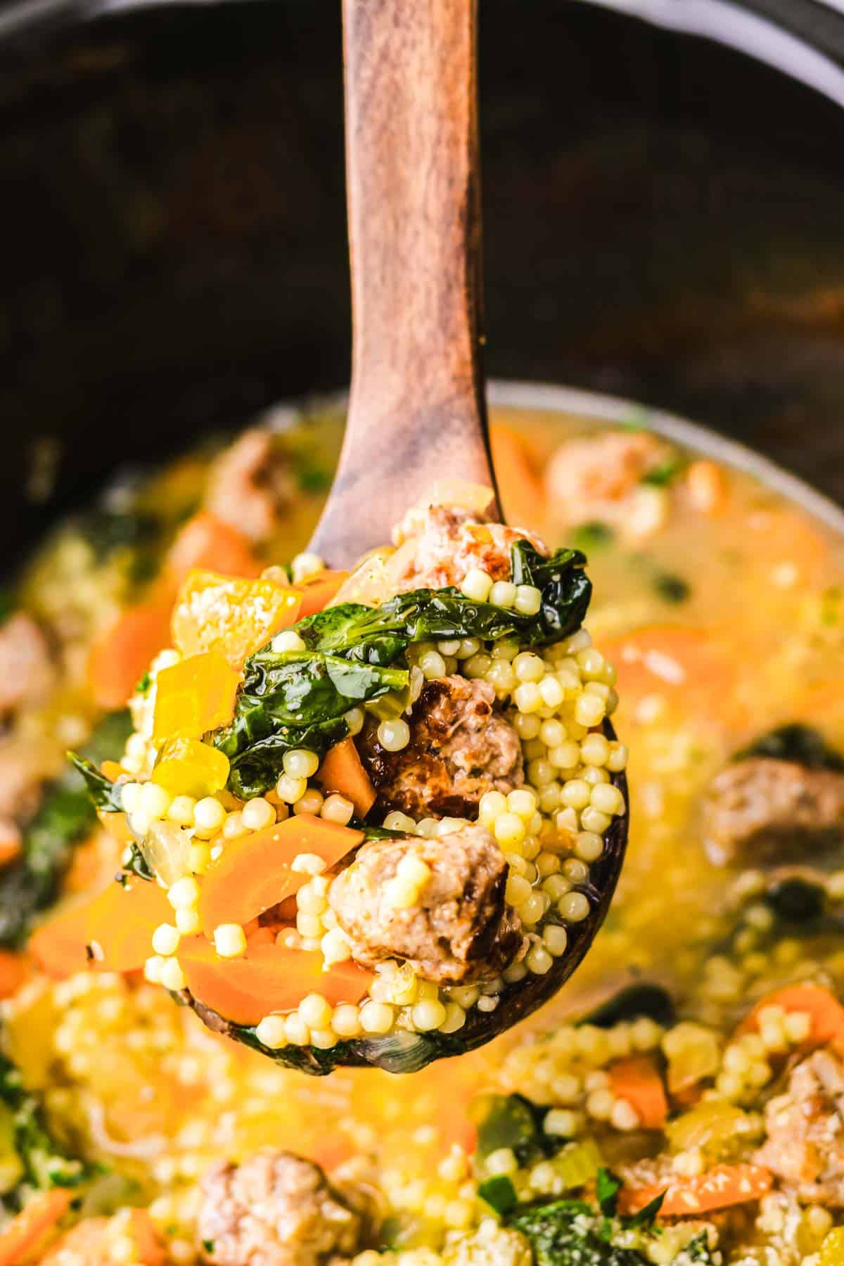 A wooden spoon holds Slow Cooker Italian Wedding Soup with sausage, couscous, kale, and carrots above a pot.